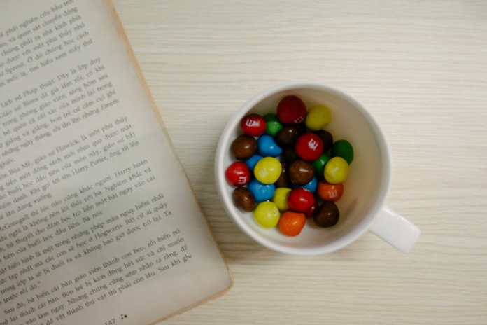 An open book and a white mug of colorful M&M's on a light wooden table, suggesting a rewarding and mindful study session.