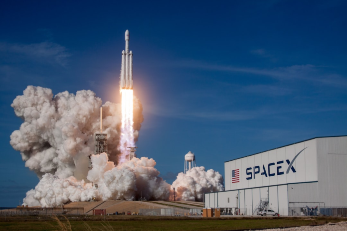Powerful SpaceX Falcon Heavy rocket launches with brilliant orange exhaust and billowing white smoke from its pad, reaching skyward under a clear blue sky, embodying the pioneering spirit of modern space exploration.