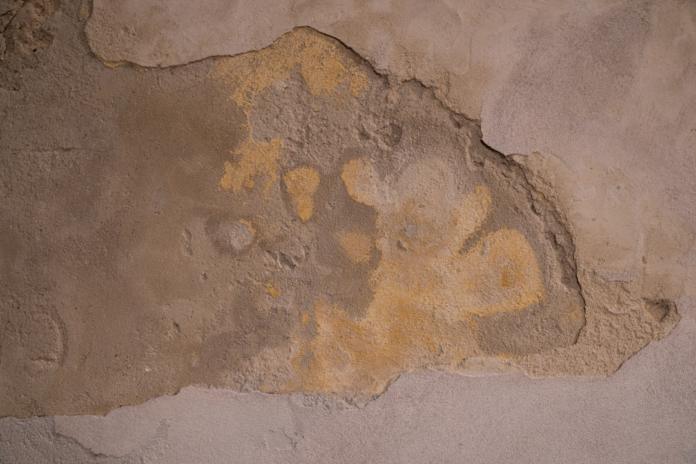 Weathered wall with peeling plaster exposing deep, textured layers of tan and ochre, evoking the unearthing of ancient history or geological strata.
