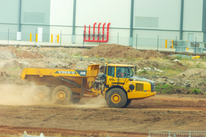 A large yellow Volvo A30E articulated dump truck actively moves across a dusty, excavated construction site with mounds of earth and an industrial building in the background, illustrating the significant human reshaping of the planet's surface.