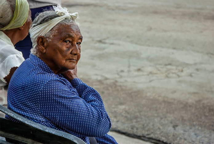 Elderly woman with a white headscarf and blue polka-dotted shirt sits on a bench, her thoughtful gaze reflecting human observation against a blurred concrete backdrop.