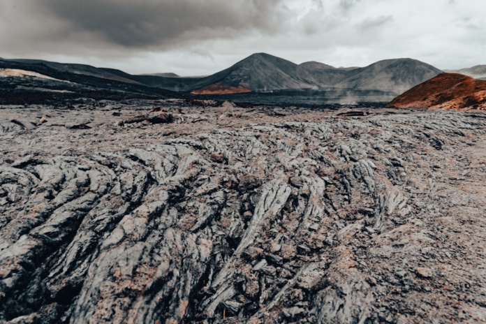 Expansive grey igneous rock formations, characteristic of cooled lava flows, dominate the foreground of a rugged volcanic landscape featuring dark mountains, reddish earth, and wisps of steam under a dramatic overcast sky.