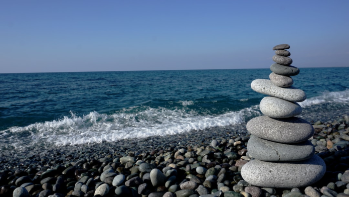 Balanced gray stones stacked on a pebble beach beside the blue ocean and clear sky, symbolizing the Earth's crust's gravitational balance and geological isostasy.