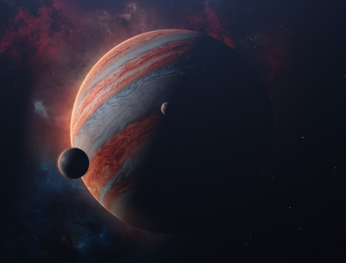 Massive, banded Jupiter is dramatically illuminated alongside two of its moons, set against a vibrant deep space nebula and distant stars.