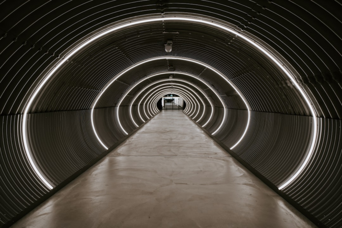 A long, futuristic tunnel with repeating bright arched lights, resembling a particle accelerator like the LHC, conveying a sense of deep scientific inquiry and the mysteries of black hole research.