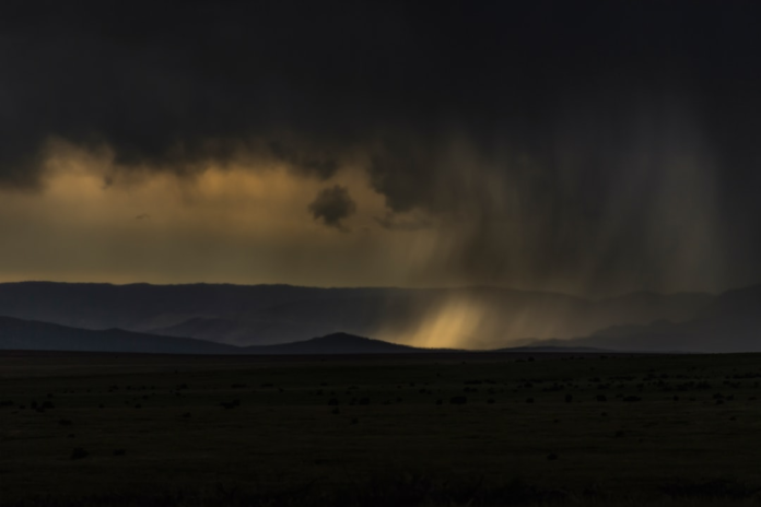 A powerful rainstorm creates dramatic light shafts over dark, sprawling plains and silhouetted mountains under a heavy, foreboding sky, symbolizing intense global weather events.