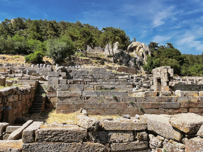 Weathered ancient stone ruins, composed of meticulously cut lithic blocks and steps, sprawl across a sunlit, tree-covered hillside with natural geological formations, reflecting the deep history of stone use in ancient landscapes.