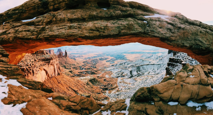 The underside of a massive natural sandstone arch glows vibrant orange from the rising sun, framing a sprawling, snow-dusted desert canyon filled with striking geological formations.