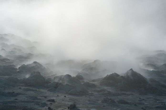 Steaming, dark basaltic rock formations emerge from a dense, ethereal geothermal fog in a rugged volcanic landscape.