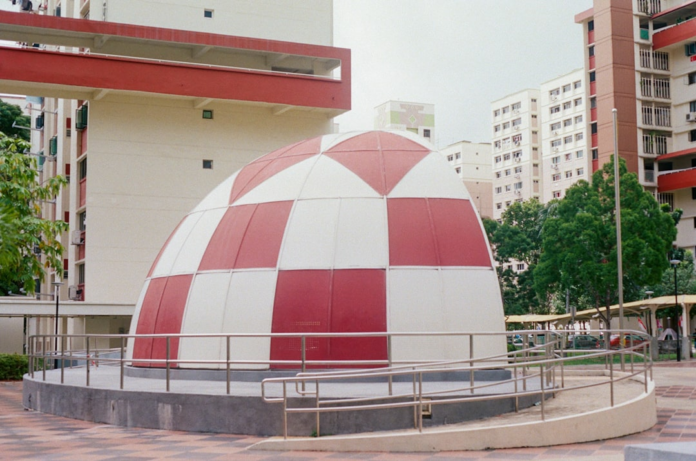 A striking red and white checkered dome, resembling a Martian habitat prototype, stands in a bustling urban landscape of towering residential buildings and green foliage, contrasting Earth's developed spaces with the challenges of settling the Red Planet.