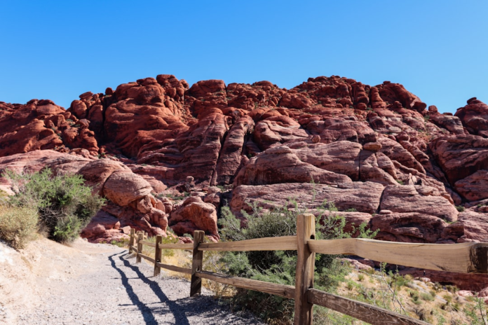 Vibrant red rock formations dominate a sunlit desert landscape with a dirt path and rustic wooden fence, evoking the rugged Martian environment for future sample return missions.