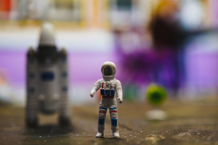 A small astronaut toy stands prominently on a dark, textured surface, with a blurred robot figure and vibrant, abstract space scenery in the background, evoking the dreams of future Mars exploration.