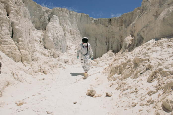 A lone astronaut in a reflective silver spacesuit traverses a rugged, white, Mars-like landscape under a clear blue sky, symbolizing future planetary exploration.