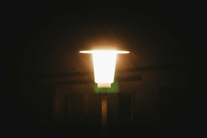 A solitary, bright lamp illuminates falling rain against a deep, dark backdrop, suggesting a moment of clarity amidst universal mysteries.