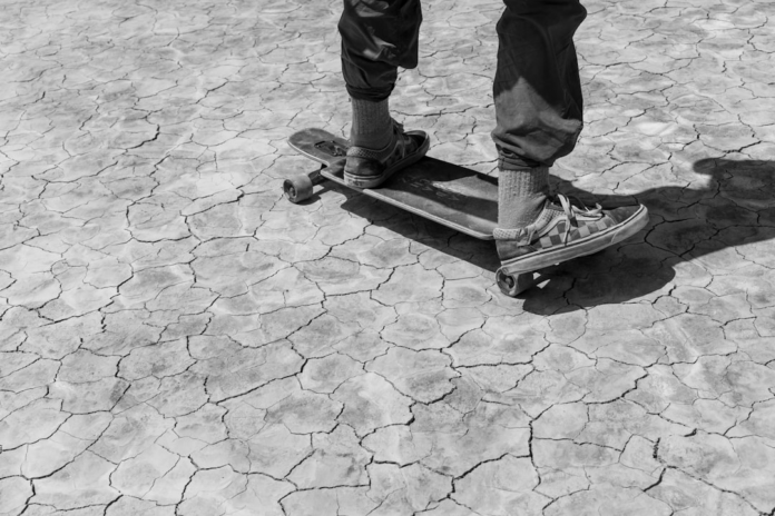 Black and white shot of a person on a longboard traversing a vast, deeply cracked dry lakebed, highlighting geological patterns and the impact of Earth's dynamics.