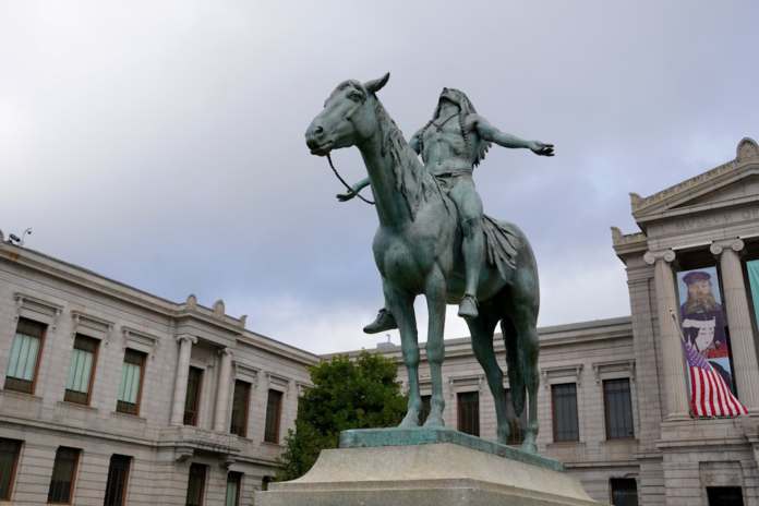 Dignified bronze statue of a Native American man on horseback, looking skyward with outstretched arms, stands prominently before a grand neoclassical museum building under an overcast sky.