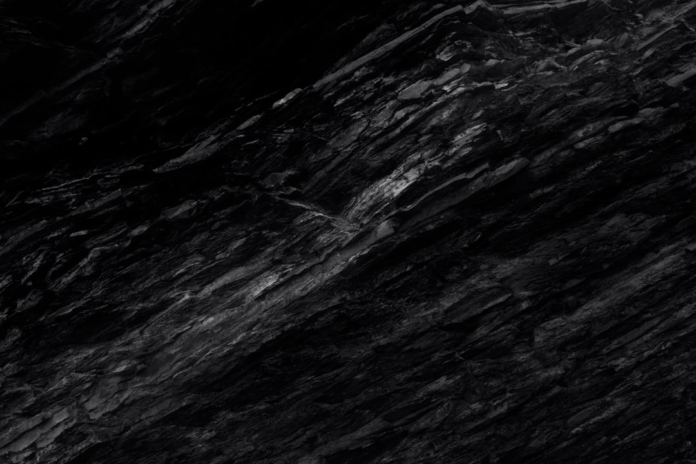 Textured dark gray rock face with distinct diagonal layers and rugged crevices, symbolizing the deep time and powerful forces explored in geology.