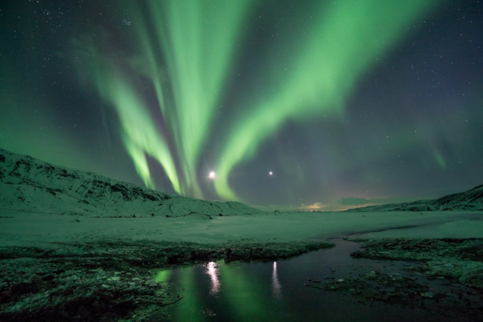 Vibrant green Northern Lights dramatically illuminate a vast, star-filled night sky and reflect in a dark, icy stream, set against a backdrop of snow-covered mountains, conveying an awe-inspiring cosmic connection.