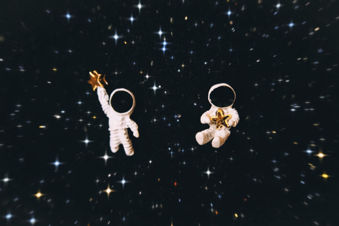 Two astronaut figurines, one reaching and one cradling a golden star, float in the vastness of deep space amidst a luminous field of twinkling stars, symbolizing human exploration.