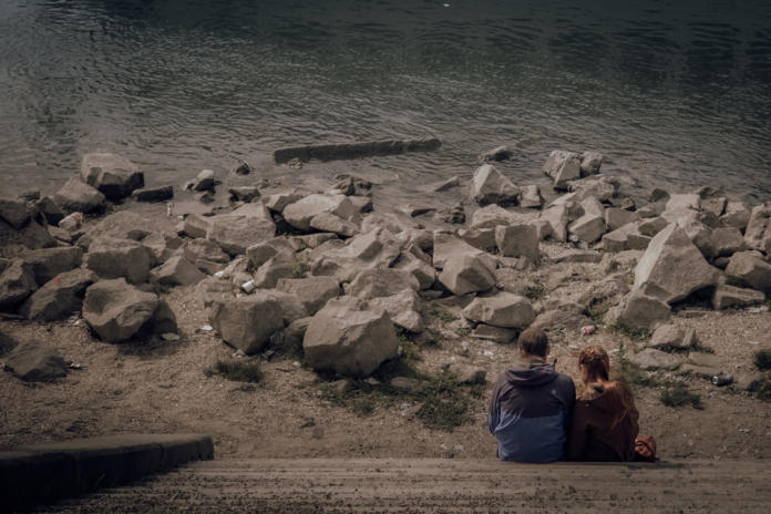 Two people sit on concrete steps overlooking a polluted riverbank, where dark water meets a rocky shore visibly strewn with plastic bottles, cans, and debris, highlighting widespread environmental degradation.