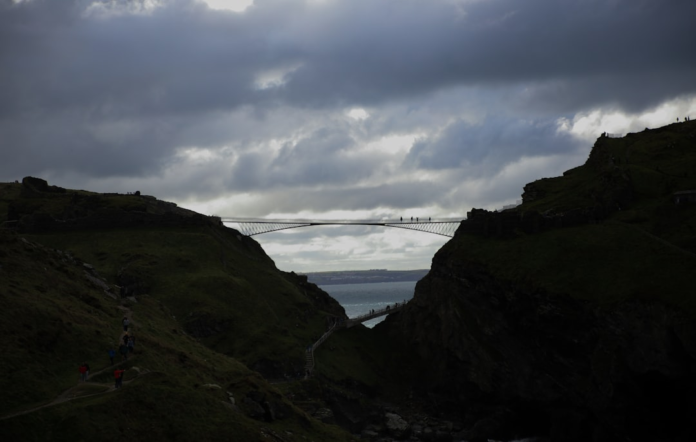 Silhouetted figures traverse a modern bridge connecting dramatic, dark cliffs above a turbulent sea under a foreboding sky, embodying the challenging path of evolution and transition.