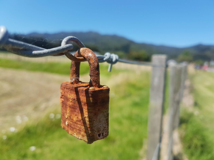 A heavily rusted padlock hangs on a wire fence under a bright sky, symbolizing a compromised barrier and the urgent need for robust privacy and data security protection.