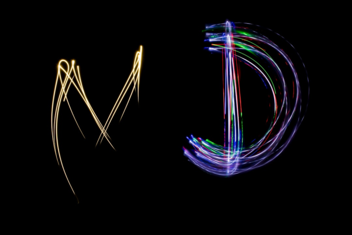 Two vibrant light trails, one golden and flowing abstractly, the other multicolored and clearly shaped like a 'D', glow mysteriously against a deep black background, hinting at the complex, interconnected nature of quantum phenomena.