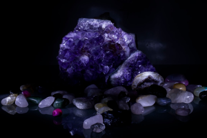 Radiant purple amethyst geode and a diverse assortment of polished quartz and other mineral stones are beautifully illuminated against a deep black, reflective background, showcasing their natural geological beauty.