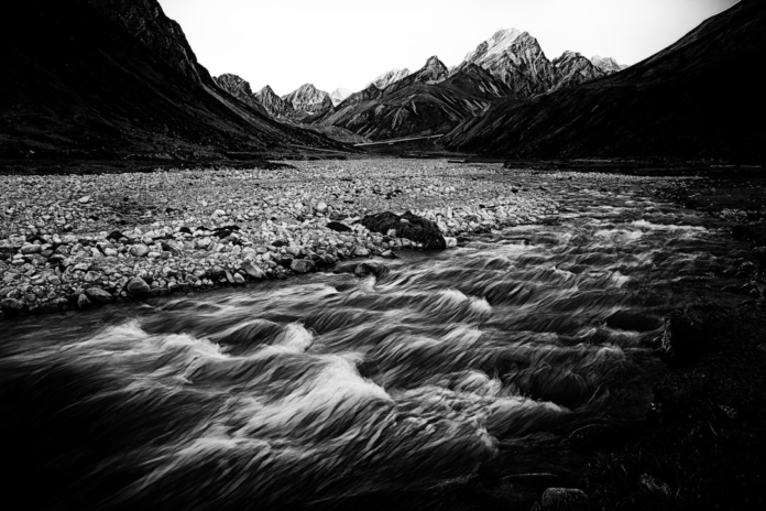 Dramatic black and white long exposure shows a powerful river flowing over a wide, rocky riverbed, framed by towering mountains, visually representing active landscape shaping and sediment transport.