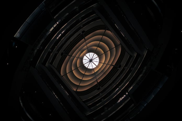 A dramatic low-angle view inside a dark, spiraling structure, featuring concentric illuminated rings converging on a bright central light, powerfully evoking a black hole's stellar accretion disk.