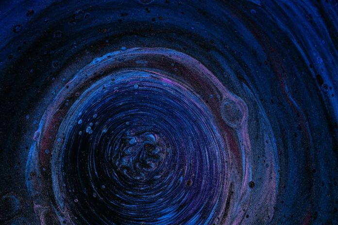 Abstract depiction of a deep blue and purple cosmic vortex, swirling with speckled light and dark bubbles, reminiscent of a supermassive black hole's accretion disk.