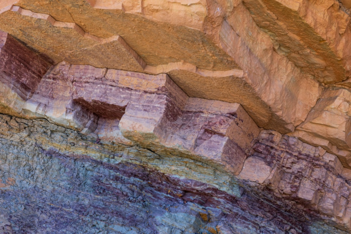 Close-up of layered geological rock with distinct bands of tan, reddish-purple, and blue-grey, showcasing the natural stratification and potential cleavage planes characteristic of slate formation.