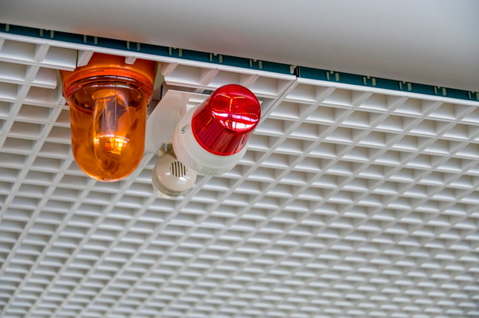 Bright orange and red security strobe lights and a small sensor are mounted on a white grid ceiling, symbolizing an alert system crucial for detecting and preventing social engineering attacks like phishing.