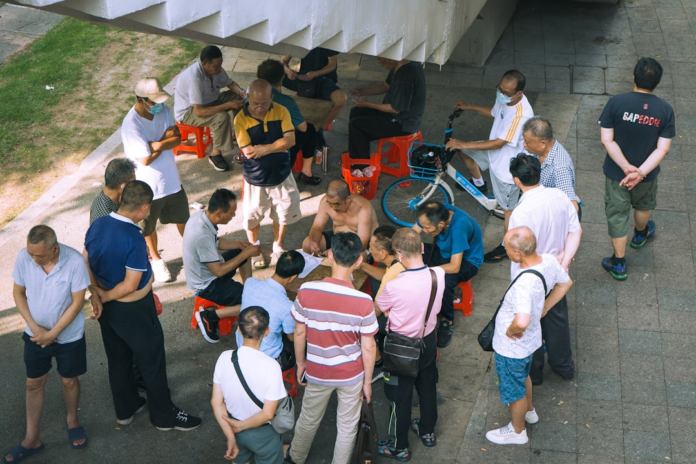 An overhead view captures a gathering of primarily older Asian men engaged in conversation and a game on a paved outdoor area shaded by a concrete structure, exemplifying observational learning and community bonding.