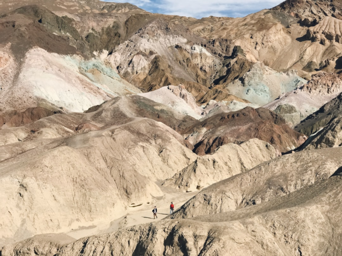 Expansive multicolored geological formations and textured sediment layers stretch across an arid desert landscape, with two small hikers in the foreground emphasizing the immense scale of Earth's ancient past.