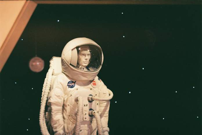 White NASA astronaut space suit, worn by a mannequin with a clear visor, is displayed against a dark, star-filled background, reflecting the historic Apollo-Soyuz era of space exploration.