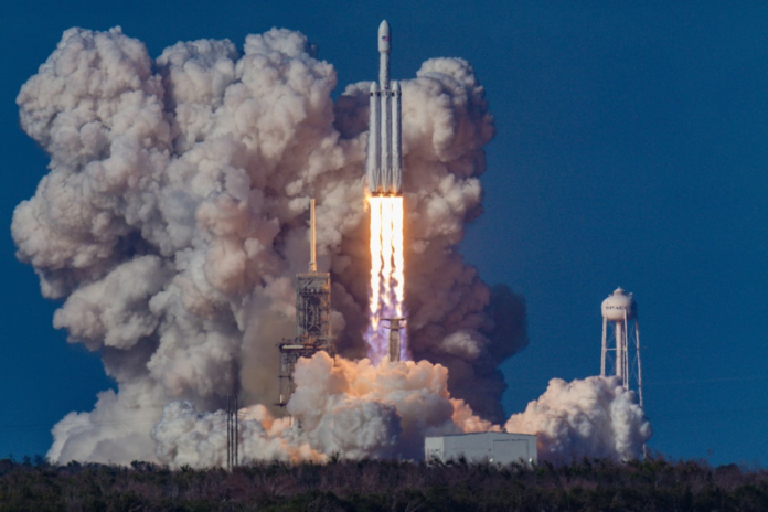 Powerful SpaceX Falcon Heavy rocket launching with fiery exhaust and immense smoke plumes from its launchpad, under a clear blue sky, signifying advanced commercial space travel capabilities near a visible SpaceX water tower.