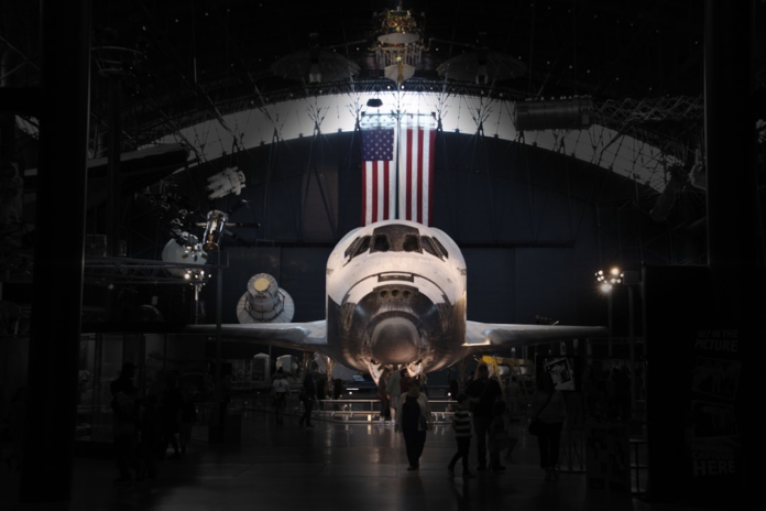 The Space Shuttle Columbia stands prominently, nose-forward, in a vast, dimly lit museum hall, illuminated beneath a large American flag as visitors observe the historic craft.