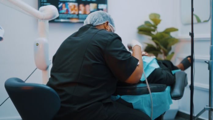 A specialized dental professional, wearing scrubs, a hairnet, and mask, leans over a patient in a modern dental chair, diligently performing an oral health procedure within a clean clinic environment.