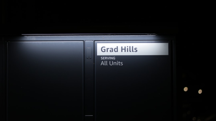 An illuminated sign on a dark, modern delivery locker displays Grad Hills and SERVING All Units, signifying an automated last-mile logistics destination.