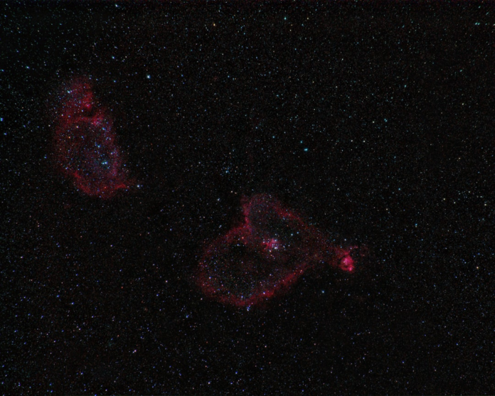 The vibrant Heart and Soul Nebulae glow in reddish hues against a dark, star-filled cosmic expanse, symbolizing the vast, ever-evolving structures of the universe.