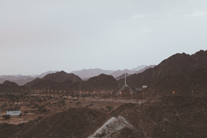Rugged, dark desert mountains loom over an expansive arid valley with a winding road, scattered streetlights, and sparse vegetation under a pale, muted sky, showcasing complex surficial geological formations.