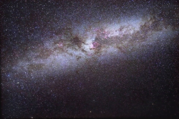 The luminous, star-studded band of the Milky Way galaxy, interspersed with dark dust clouds and faint pink nebulae, glows majestically across the deep cosmic night.