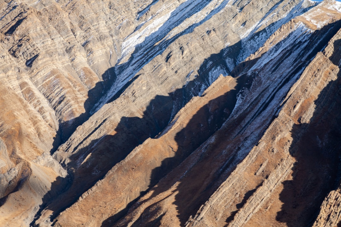 Rugged, stratified mountain slopes display distinct geological rock layers, with dramatic shadows and sparse snow highlighting the steep, eroding terrain prone to rockfalls.