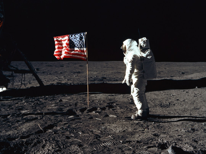 A solitary astronaut in a bright white spacesuit stands next to a planted American flag on the desolate grey lunar surface under a dark sky, powerfully symbolizing humanity's pioneering spirit in space exploration.