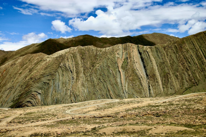 Expansive mountain landscape featuring impressive folded and stratified geological rock formations, traversed by a winding dirt road under a partially cloudy blue sky, conveying Earth's ancient dynamics.