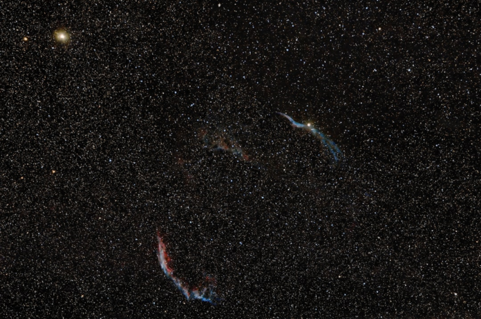 Vast cosmic expanse showing the luminous, arcing filaments of the Cygnus Loop supernova remnant in blue and red, with a bright star and countless other stars scattered across the dark background, highlighting universal change.