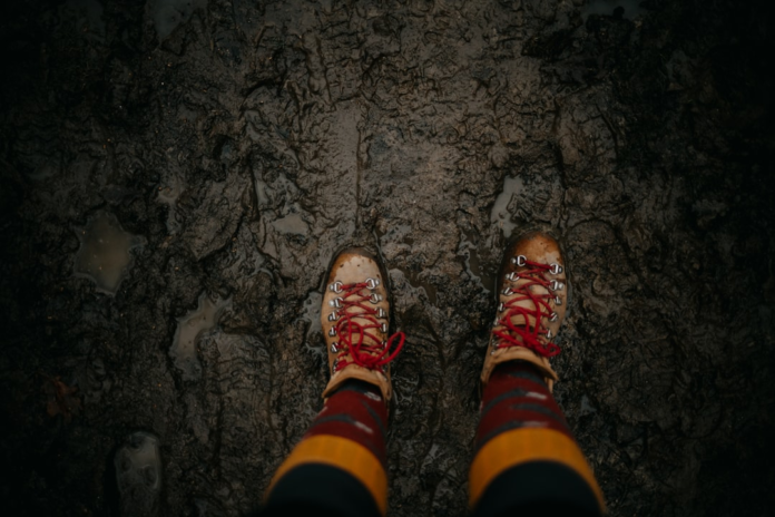 Brown hiking boots with vibrant red laces stand firmly in thick, dark mud on a rugged trail, symbolizing the vigorous outdoor activity essential for improving VO2 Max and promoting longevity.
