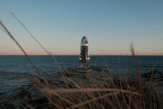 A gleaming silver rocket statue, symbolizing pioneering space exploration like the Voskhod program, stands on a rocky coastal outcrop overlooking a vast blue ocean, framed by golden foreground grasses under a serene twilight sky.