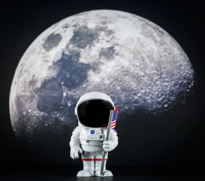 A detailed astronaut figurine in a white suit holds an American flag, set against a large, illuminated cratered moon, symbolizing the ambition of Apollo era moon journeys.
